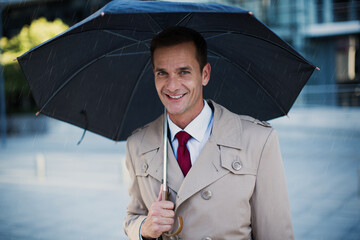 Smiling businessman in trench coat under umbrella