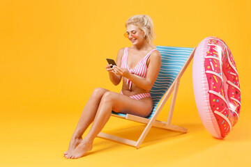 Smiling young woman girl in pink striped swimsuit glasses sit on deck chair isolated on yellow background studio. People summer vacation rest lifestyle concept. Using mobile phone, typing sms message.
