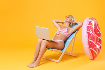 Smiling young woman in pink striped swimsuit glasses sit on deck chair isolated on yellow background. People summer vacation rest lifestyle concept. Working on laptop computer hold hands behind head.