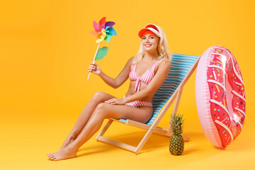 Smiling young woman girl in pink striped swimsuit cap sit on deck chair isolated on yellow wall background studio. People summer vacation rest lifestyle concept. Mock up copy space. Hold toy windmill.