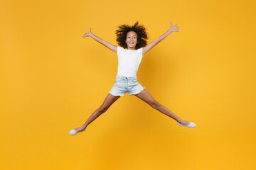 Fototapeta na wymiar Full length portrait of excited little african american kid girl 12-13 years old in white t-shirt isolated on yellow wall background. Childhood lifestyle concept. Jumping, spreading hands and legs.