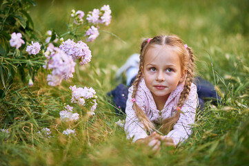 The little girl lies on the green grass near the bush with pink flowers and makes different facial expressions. girl in pink blouse and blond hair