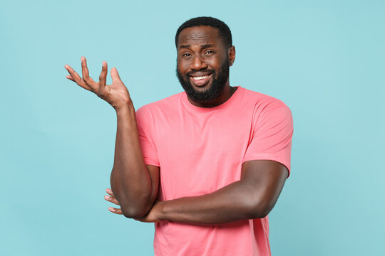 Confused smirked young african american man guy in casual pink t-shirt isolated on blue wall background studio portrait. People sincere emotions lifestyle concept. Mock up copy space. Spreading hands.