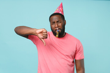 Displeased young african american man guy in casual pink t-shirt birthday hat isolated on pastel blue wall background. People lifestyle concept. Mock up copy space. Blowing in pipe showing thumb down.