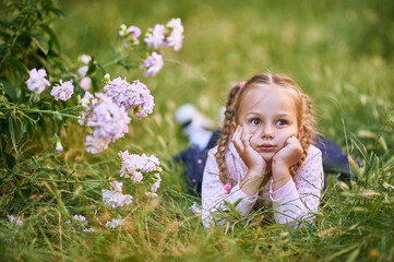 The little girl lies on the green grass near the bush with pink flowers and makes different facial expressions. girl in pink blouse and blond hair