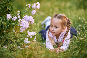 The little girl lies on the green grass near the bush with pink flowers and makes different facial expressions. girl in pink blouse and blond hair