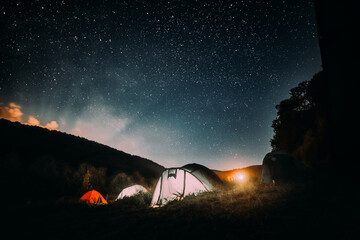 Summer camping in the mountains. Tents in the night with the starry sky and clouds in the background. Illuminated tents on the grassy ground in the mountains.