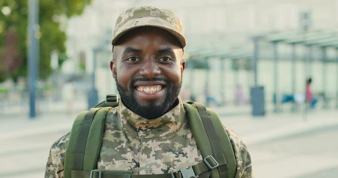 Portrait of cheerful African American handsome young male soldier in cap with backpack smiling at street. Close up of male militarian with happy smile at train station. Bus stop. Military uniform.