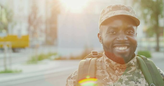 Portrait of joyful handsome African American young male soldier in cap and with backpack smiling to camera at street. Close up of man militarian with happy smile in city outdoors. Military uniform.