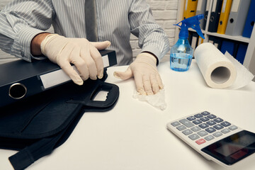 concept of cleaning or disinfecting the office desk - a businessman cleans the workplace, computer keyboard, document folders, uses a spray gun sanitizer, gloves and paper napkins.