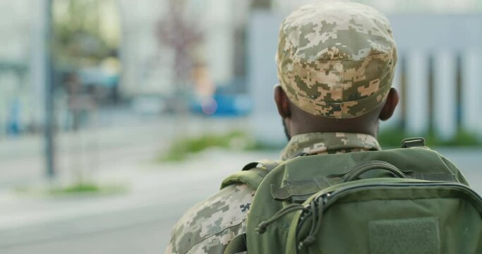 Rear of African American young male soldier in uniform, hat and with backpack walking outdoor. Back view on militarian officer strolling the street in military clothes. Going to army.