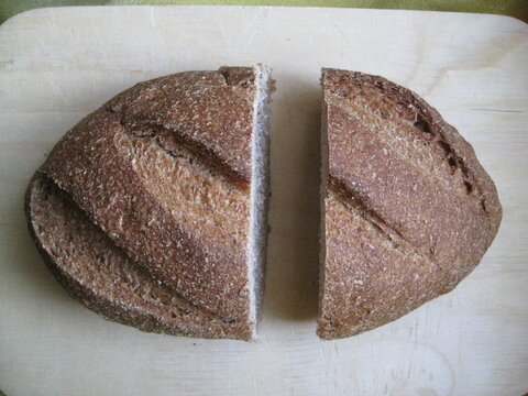 pan de levadura madre, hogaza de forma oval con tres grietas de cocci&oacute;n cortada en dos pedazos separados sobre tabla madera.
