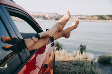 Female legs hanging out of a car window, and a hand holding sunglasses, by the lake. Concept of travel and summer vacation.