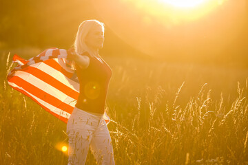 Beautiful young girl holding an American flag in the wind in a field of rye. Summer landscape against the blue sky. Horizontal orientation.