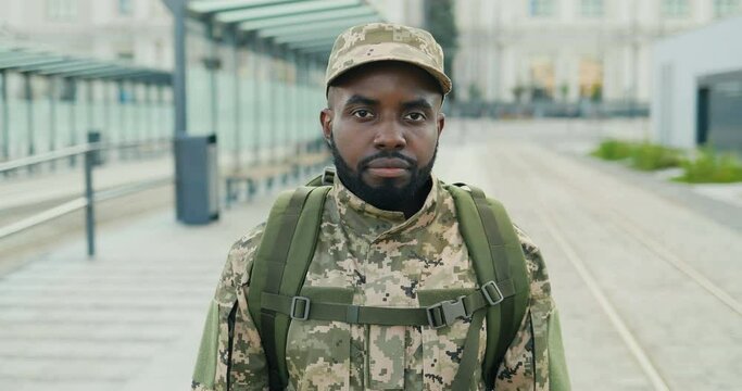 Portrait of serious African American handsome young male soldier in cap with backpack standing at street. Man militarian looking at camera at train station. Bus stop. Military uniform. Zooming in.
