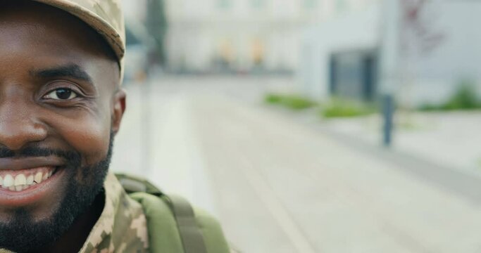 Portrait of cheerful African American handsome young male soldier in cap with backpack smiling at street. Close up of half face of man militarian with happy smile outdoors. Military uniform.