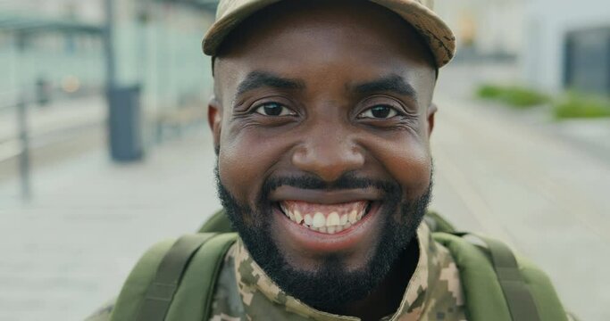 Portrait of cheerful African American handsome young male soldier in cap with backpack smiling at street. Close up of man militarian with happy joyful smile outdoorn in town. Military uniform.