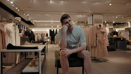 A bored man sits on a bench in a clothing store. He is tired of waiting for his girlfriend to choose a fashionable outfit.