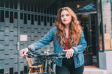 Fototapeta na wymiar Red curled long hair caucasian teen girl on the city street walking with bicycle fashion portrait. Natural people beauty urban life concept image.