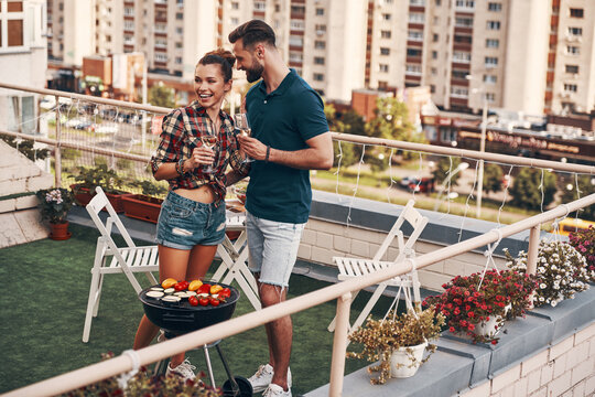 Good looking young couple in casual clothing preparing barbecue and smiling while standing on the rooftop patio outdoors