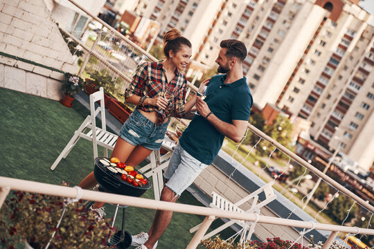 Charming young couple in casual clothing preparing barbecue and smiling while standing on the rooftop patio outdoors
