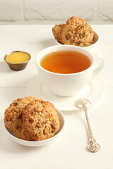 Oatmeal cookies with walnuts and raisins on small saucers and a cup of green tea, standing on a white wooden table against a white brick wall. Closeup