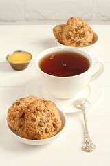 Oatmeal cookies with walnuts and raisins on small saucers and a cup of black tea, standing on a white wooden table against a white brick wall. Closeup