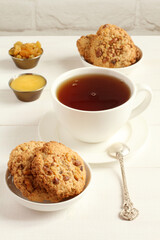 Oatmeal cookies with walnuts and raisins on small saucers and a cup of black tea, standing on a white wooden table against a white brick wall. Closeup