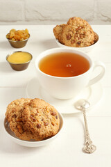 Oatmeal cookies with walnuts and raisins on small saucers and a cup of green tea, standing on a white wooden table against a white brick wall. Closeup