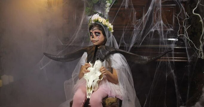 Portrait of girl dressed in Halloween Santa muerte costume  holding a goat scull against background of old dark Gothic interior covered with cobwebs and mysterious fog. 4K slow motion 50 fps