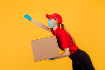 agitated young delivery woman, in a red uniform, wearing a medical protective mask and gloves, running and holding a cardboard box isolated on a yellow background. Express delivery, delivery service