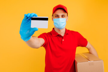 Obraz na płótnie Canvas Courier-a man in a medical mask and gloves, in a red cap, t-shirt, uniform. A delivery man holds a cardboard box and shows a credit card. Online stores, delivery service, coronavirus