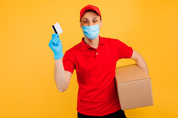 Fototapeta na wymiar Courier-a man in a medical mask and gloves, in a red cap, t-shirt, uniform. A delivery man holds a cardboard box and shows a credit card. Online stores, delivery service