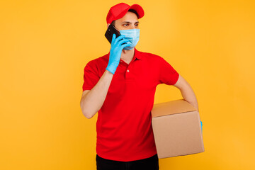 Fototapeta na wymiar male courier in a red uniform, medical mask and gloves holds a cardboard box and talks on a mobile phone on a yellow background. Delivery service, online stores