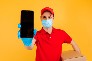 Fototapeta na wymiar male courier in a red uniform, medical mask and gloves holds a cardboard box and shows an empty mobile phone screen on a yellow background. Delivery service, online stores, coronavirus