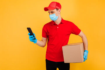Fototapeta na wymiar male courier in a red uniform, medical mask and gloves holds a cardboard box and uses a mobile phone on a yellow background. Delivery service, online stores