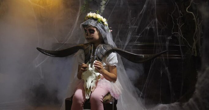 Portrait of girl dressed in Halloween Santa muerte costume  holding a goat scull in of old dark Gothic interior covered with cobwebs and mysterious fog and  changing lighting. 4K slow motion 50 fps
