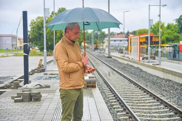 Portrait of a mature man with an umbrella standing on the platform of a train station. Unfinished railway line. When the train arrives.. Funny concept of hopeless situation