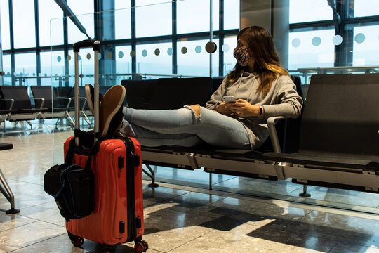 Attractive young female waiting at the airport to board a flight with her legs stand on top of a red hand luggage
