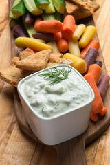 Tzatziki. Traditional Greek or Turkish homemade dip. Made with Greek yogurt, cucumbers, lemon juice, olive oil and dill. Served with mini rainbow carrots & crispy pita chips on a wooden cutting board
