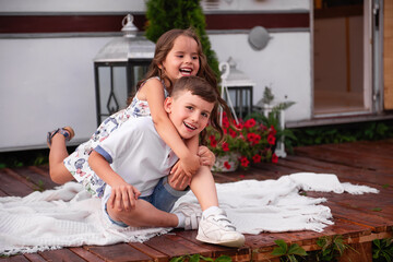 Little beautiful girl hugs the boy from behind. Brother and sister laugh, smile, play at their trailer, mobile home. Happy family travels with children on summer vacation. Summer picnic camp concept