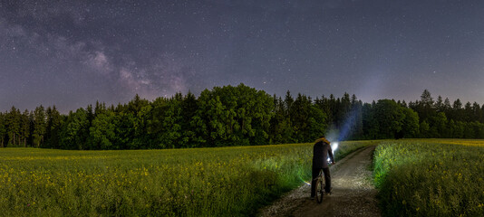 Panoramic photo of a man with his bike watching the beauty of the milkyway at the dark night sky at...