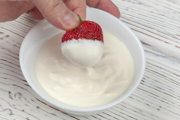 men's hand dips strawberries in sour cream on white wooden background