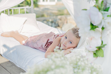 Obraz na płótnie Canvas Close up portrait blonde girl with long hair in a beautiful pink powdery dress sleep in a white bed with chiffon canopies, decorated with wisteria, flowers against the background of the sea. Magic day