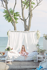 Delicate little girl blonde with long hair in a beautiful pink powdery dress sits by a white bed with chiffon canopies, decorated with wisteria, flowers against the background of the sea. Design 