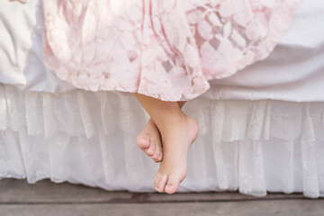 A little blonde girl in a delicate pink dress sits on a white bed with her bare feet dangling down....