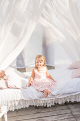 Fototapeta na wymiar A little blonde girl in a delicate pink dress sits on a white bed with her bare feet dangling down. The photo zone is decorated with wisteria flowers, chiffon canopies, a birthday photo session