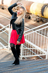 A beautiful, modern, fashionable blonde girl with long hair, red lipstick stands  on the steps. Dressed in a skirt, black leather jacket, boots, tights. Young mother. A fit figure, a respectable face