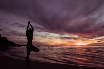 Silhouette of young woman wearing exercise clothes practicing yoga on the beach at sunset or sunrise. women do yoga. women exercising yoga in the beach. Concept : Health care
