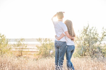 Naklejka na ściany i meble Beautiful couple stand in the dry grass with their backs in jeans, watch the sunset. Young red-haired man hugs a brunette slender girl. Belief in a good future, a free life, travel hitchhiking for two